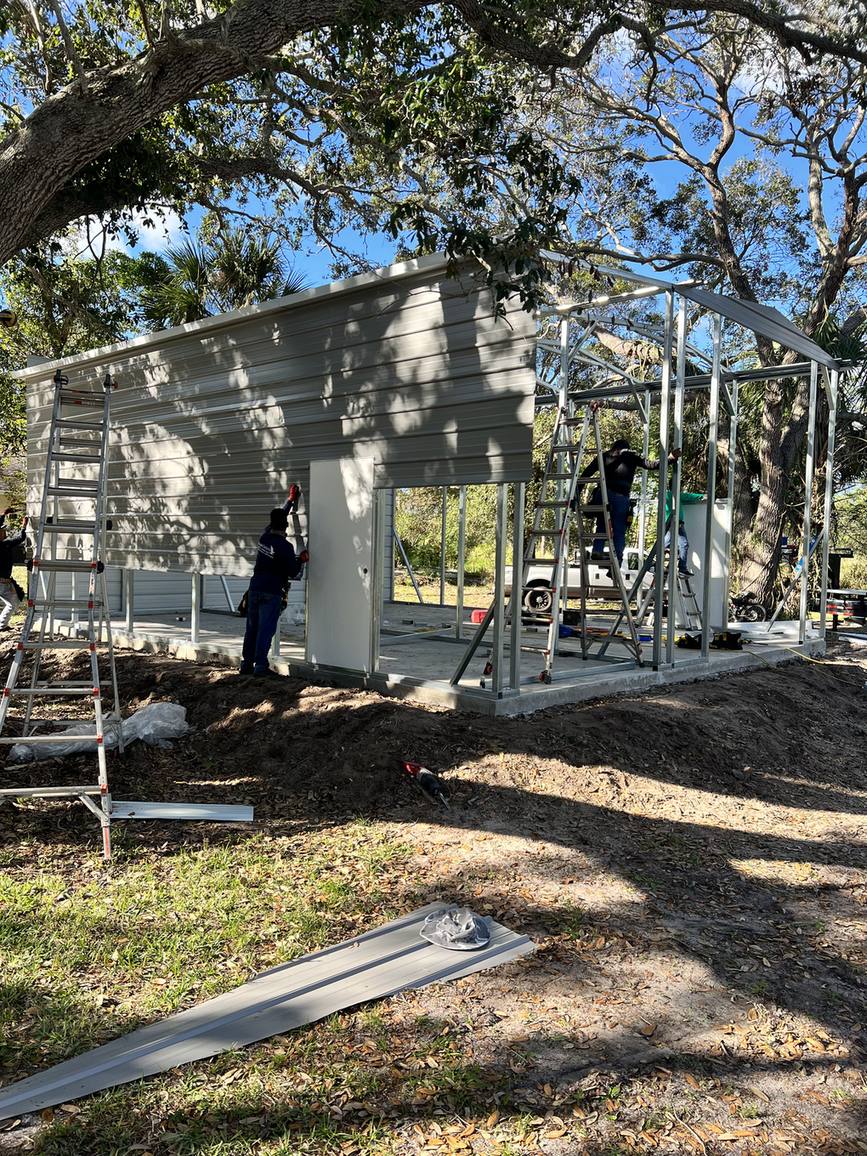 Crew fastening wall panels on a metal garage under live oaks